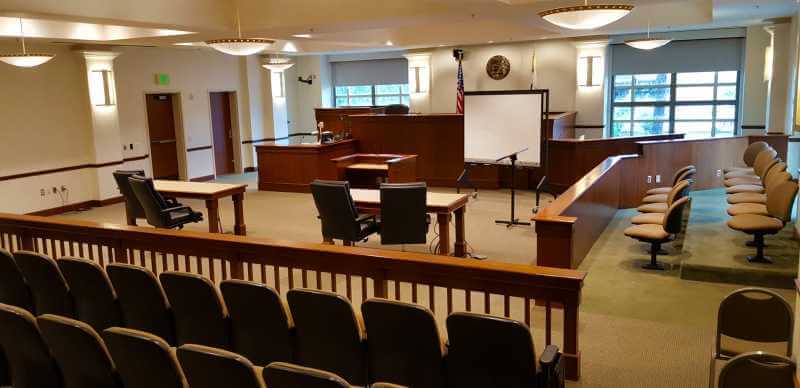 This is a photo of inside a courtroom. The photo shows the jury box to the right; counsel tables; judges bench; witness box; clerks box and the gallery. It also includes a screen.